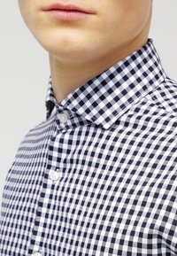 Camicia a quadretti vichy blu navy e bianca con colletto abbottonato, caratterizzata da una texture liscia e bottoni bianchi lungo la patta.