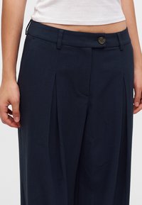 Gros plan d'une personne portant un pantalon large bleu marine avec des plis sur le devant et une ceinture boutonnée, associé à un top blanc court.