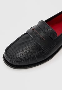 Sorte læder loafers med et struktureret reptilmønster, rød indvendig foring og en dekorativ rem med udskæringer på overdelen.