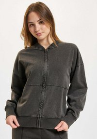 Tamno siva dukserica na zip s teksturiranim materijalom, rebrastim manžetama i rubom, s vidljivim detaljima šivanja i ležernim krojem.