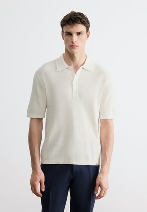 Nuori mies, joka pukeutunut kermaiseen lyhythihaiseen neulottuun poolopaitaan ja tummansinisiin housuihin, seisoo yksivärisen valkoisen taustan edessä.