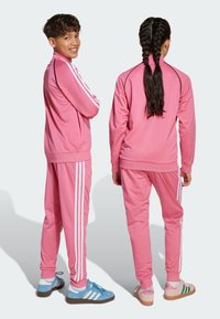 Ružové teplákové súpravy s bielymi pruhmi, ktoré majú zipsové bundy a elastické manžety. Tenisky v rôznych farbách dokončujú športový vzhľad.