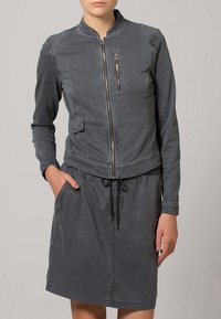 Chaqueta vaquera gris con cremallera frontal, bolsillo pequeño y puños acanalados, combinada con un vestido gris a juego con cintura ajustable con cordón.