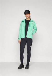 Kerge mündiroheline jakk mustade detailide ja logoga, must tihedalt istuv top, mustad retuusid ja mustad spordijalatsid.