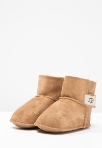 Botas para bebés de gamuza beige con forro suave, punta redondeada y una correa de Velcro con el logo de UGG. Costura visible a lo largo de los bordes.