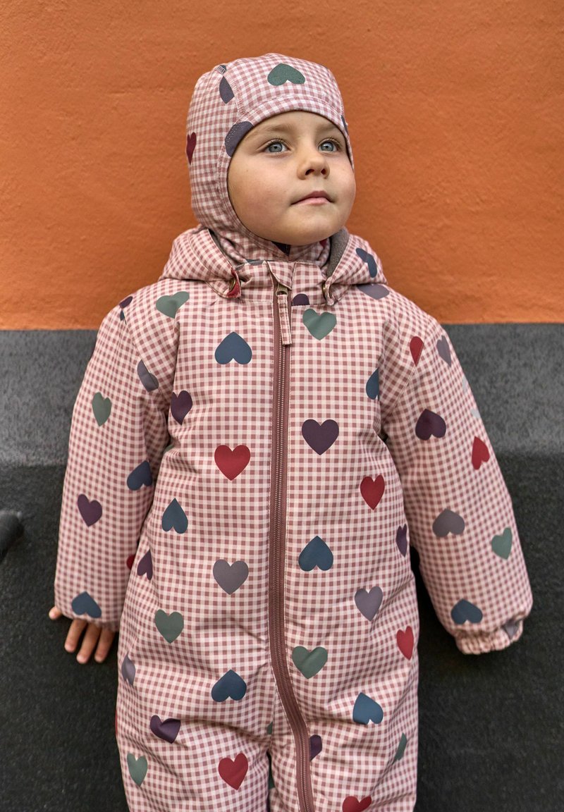 Παιδί με ροζ καρό ολόσωμη φόρμα με κουκούλα διακοσμημένη με πολύχρωμες καρδιές, στέκεται μπροστά σε πορτοκαλογκρί τοίχο.