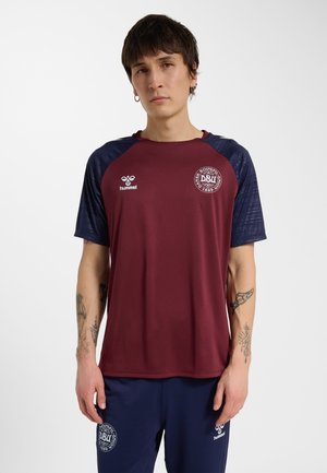 Ung mand med tatoveringer iført en vinrød og marineblå sportstrøje og matchende marineblå shorts med logoer fra Dansk Boldspil-Union.