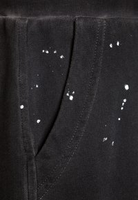 Černá látka s texturovaným povrchem, s bílými skvrnami od barvy na povrchu a detailem postranní kapsy.