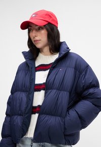 Granatowa kurtka puchowa z wysokim kołnierzem i zamkiem; noszona na białym swetrze z dzianiny w czerwone i granatowe paski; zestawiona z czerwoną czapką.