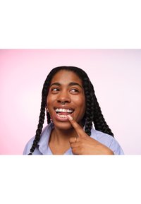 Ritratto di una persona con lunghi capelli intrecciati che indossa una camicia a righe blu, sorridente e che indica i propri denti su uno sfondo rosa.