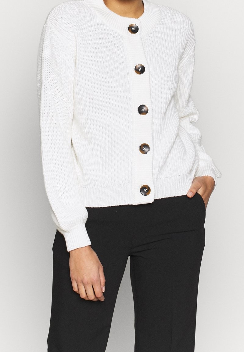 Cardigan en maille blanche boutonné avec manches longues, associé à un pantalon noir, une main dans la poche et l'autre reposant sur le côté.