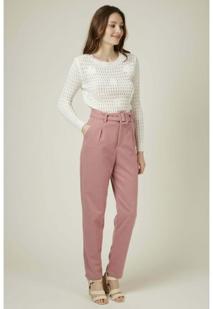 Pull en maille blanche avec un motif ajouré et des manches longues, associé à un pantalon rose taille haute avec une ceinture et des poches latérales.