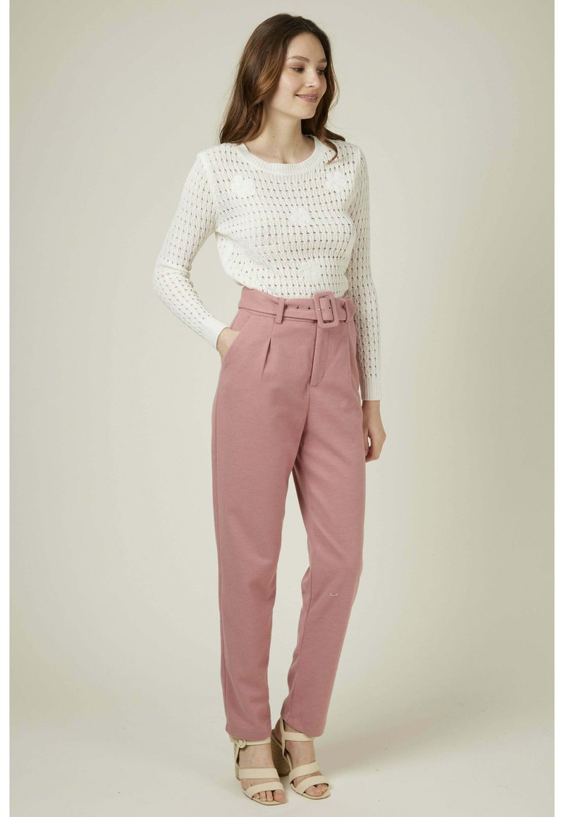 Pull en maille blanche avec un motif ajouré et des manches longues, associé à un pantalon rose taille haute avec une ceinture et des poches latérales.