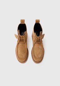 Botas de tobillo de ante marrón con cordones, que cuentan con una punta redonda, detalles de costura y una suela de goma. Incluyen una pestaña en el talón.