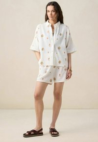 White cotton two-piece set featuring a short-sleeved button-up shirt and coordinating shorts, both with shell-pattern embroidery in gold and peach.