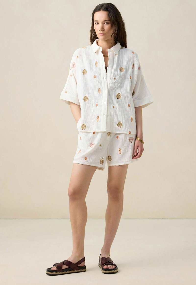 White cotton two-piece set featuring a short-sleeved button-up shirt and coordinating shorts, both with shell-pattern embroidery in gold and peach.