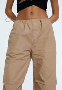 Pantalones sueltos de color beige claro, con cinturilla elástica y cordón; la tela parece suave y ligera, con una textura sutil.