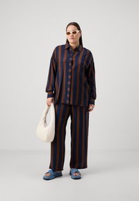 Dviejų dalių apranga, susidedanti iš didelio, rudo ir tamsiai mėlyno dryžuoto marškinio ir plačių kelnių. Priedai apima baltą rankinę ir mėlynas šlepetes.