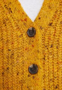 Cardigan lavorato a maglia color giallo senape con scollo a V, due bottoni rotondi scuri e un motivo testurizzato con macchie di arancione e marrone.