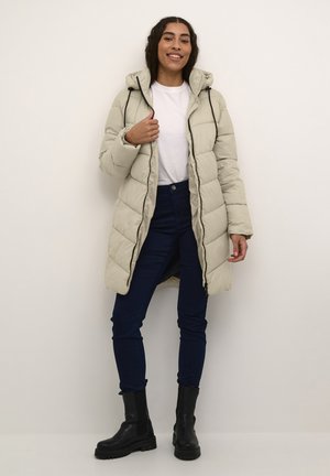 Manteau rembourré beige clair avec capuche, design matelassé, fermeture éclair à l'avant et cordon de serrage. Associé à un jean bleu marine et des bottines noires.