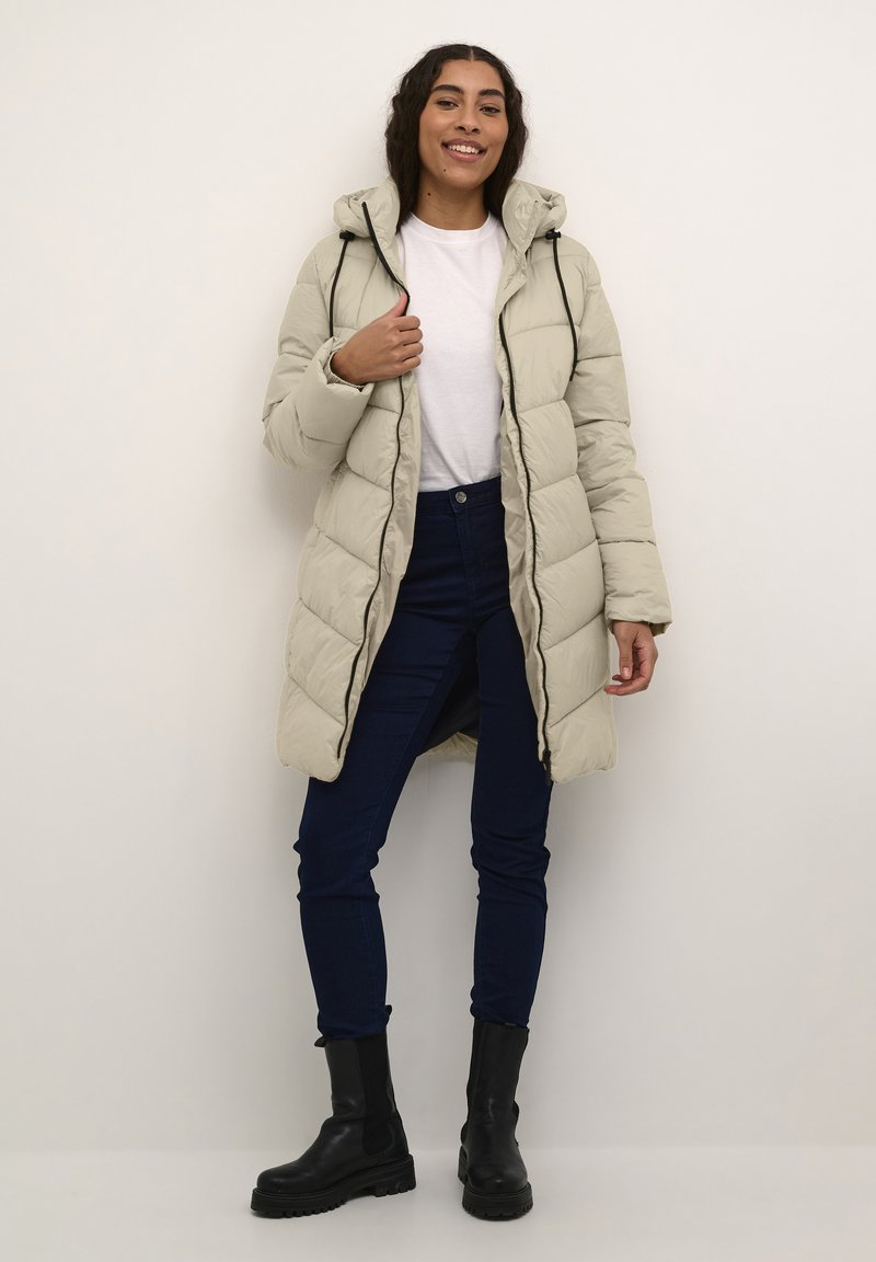 Light beige puffer coat with a hood, quilted design, front zipper, and drawstring. Paired with dark blue jeans and black ankle boots.