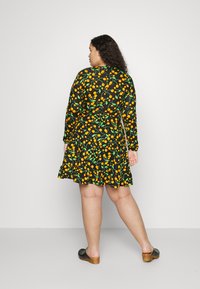 Vestido preto com mangas longas, padrão floral vibrante em laranja e verde, barra em babados, e combinado com tamancos pretos. Visto por trás.