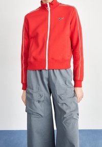 Röd zip-up munkjacka med vit kant och en svart logotyp, tillsammans med grå cargobyxor med stora fickor och avslappnad passform.