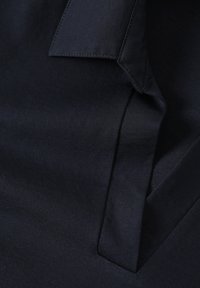 Tessuto blu navy con una superficie liscia e un leggero lucido. Presenta un design piegato e dettagli cuciti sui bordi. Dettaglio della tasca piatta visibile.