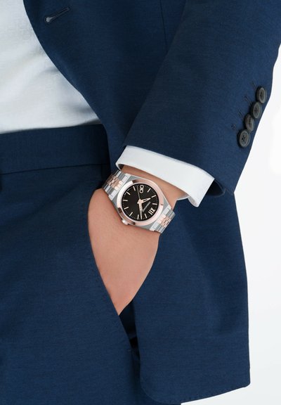 Reloj de acero inoxidable con esfera cuadrada, dial negro, acentos en oro rosa y pulsera de eslabones plateados, usado en una mano metida en el bolsillo de un traje azul marino.