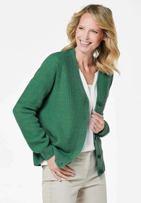 Grüne Strickjacke mit Rippstruktur, mit durchgehender Knopfleiste, langen Ärmeln und lockerer Passform. Getragen über einem weißen Oberteil.