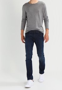 Homme portant une chemise grise à manches longues, un jean bleu foncé et des baskets blanches marchant devant un fond clair uni.