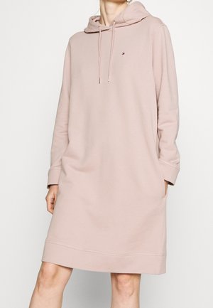 Femme portant une robe à capuche à manches longues de couleur rose clair avec des cordons de serrage et des poches latérales, debout devant un fond blanc uni.