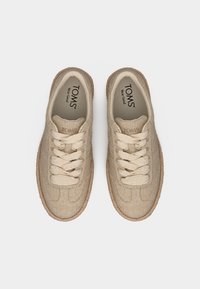 Ζευγάρι μπεζ υφασμάτινα αθλητικά παπούτσια TOMS με κορδόνια και καφέ υφή σόλες, σε θέα από ψηλά σε λευκό φόντο.