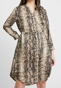 Långärmad skjortklänning i ormskinnsmönster. Har ett knytliv, knappar framtill och ett mjukt, flödande tyg. Färgpaletten inkluderar beige, svart och brun.