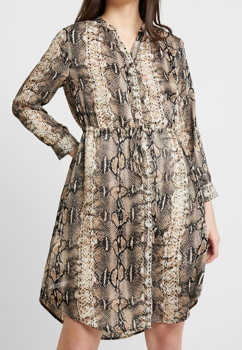 Långärmad skjortklänning i ormskinnsmönster. Har ett knytliv, knappar framtill och ett mjukt, flödande tyg. Färgpaletten inkluderar beige, svart och brun.