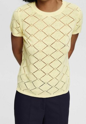 Vrouw draagt een pastelgeel gebreid topje met korte mouwen en een opengewerkt patroon in ruitvorm, gecombineerd met donkerblauwe broek.