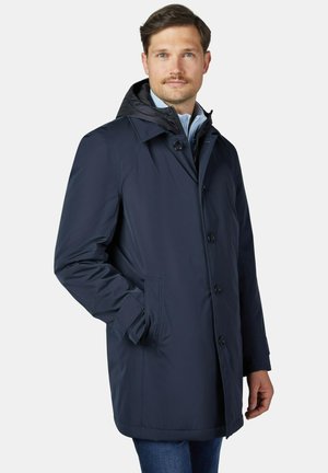Mann mit hellbraunen Haaren und Schnurrbart, der einen marineblauen Kapuzenmantel über einem hellblauen Hemd und Jeans trägt, steht mit einer Hand in der Tasche.