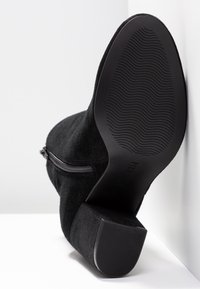 Bottine en suède noir avec fermeture éclair latérale, semelle en caoutchouc texturée, et talon bloc avec un motif strié pour une meilleure adhérence.