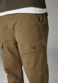 Olijfgroene cargobroek van katoen met een gladde textuur, voorzien van meerdere zakken, contrasterende stiksels en een merketiket aan de zijkant.