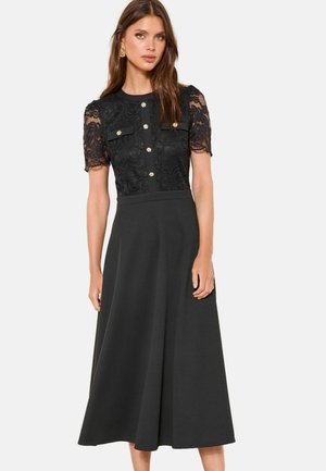 Frau in einem schwarzen Midi-Kleid mit kurzen Spitzenärmeln, dekorativen goldenen Knöpfen und zwei Brusttaschen, die vor einem schlichten Hintergrund steht.