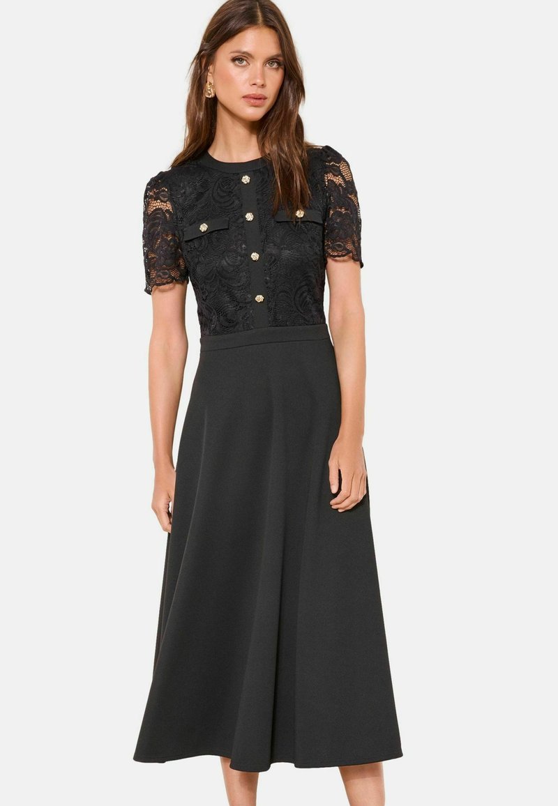 Femme portant une robe midi noire avec des manches courtes en dentelle, des boutons décoratifs dorés et deux poches poitrine, debout devant un fond uni.