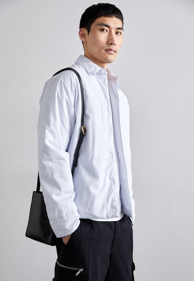 Lys blå stribet button-up skjorte med en afslappet pasform, parret med sorte bukser og en skuldertaske. Glat tekstur, synlige sømme og accenter.