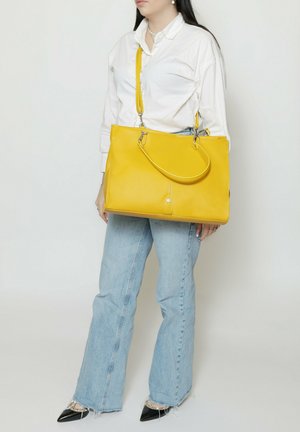 Donna con camicia bianca e jeans azzurri chiari che tiene una grande borsa gialla con tracolla su sfondo bianco.