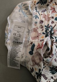 Stoffhut mit einem mehrfarbigen Tier- und Blattmuster. Hat einen weißen Hintergrund, ein elastisches Band und ein Pflegeetikett mit Anweisungen in mehreren Sprachen.