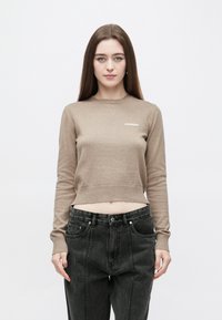 Béžový krátký svetr s dlouhými rukávy, žebrovanými manžetami a kulatým výstřihem. Na prsou je malý bílý logo. Pár černých džínových kalhot.