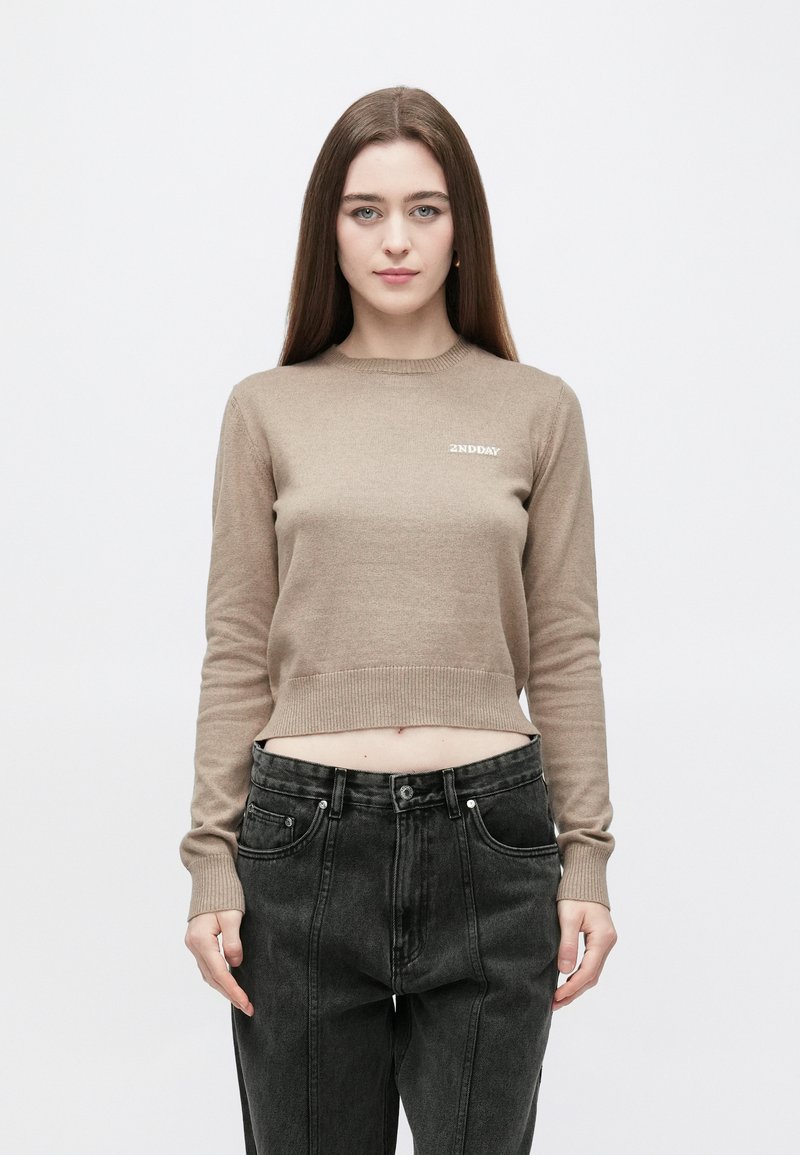 Béžový krátký svetr s dlouhými rukávy, žebrovanými manžetami a kulatým výstřihem. Na prsou je malý bílý logo. Pár černých džínových kalhot.