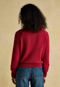 Červený pletený sveter s ribovaným lemom a manžetami, ktorý má dizajn raglánových rukávov. V kombinácii s modrými džínami s vreckami na zadnej strane.