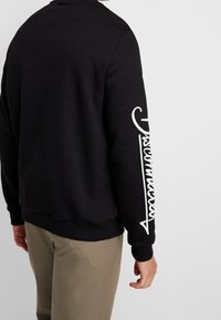 Felpa nera con polsini e orlo a costine, con lettering bianco che corre lungo la manica destra. Tessuto morbido e vestibilità comoda.