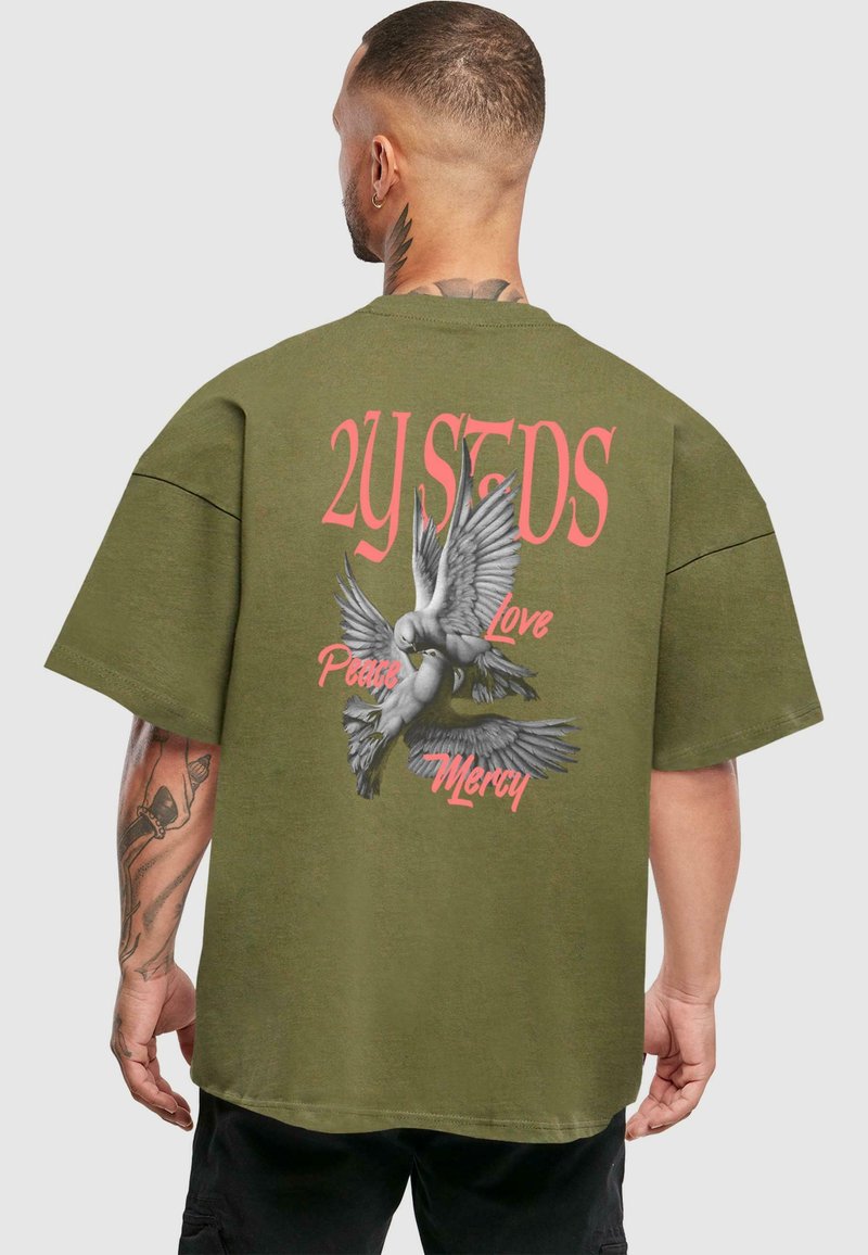 Camiseta tamaño grande en verde oliva con un estampado gráfico de palomas y el texto "2Y STDS" en rosa, con un corte holgado y mangas cortas.