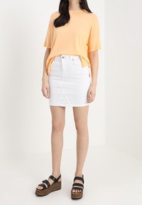Vrouw in een perzikkleurige korte mouwen top, witte denim mini rok en zwarte platform sandalen met houten gestreepte zolen.